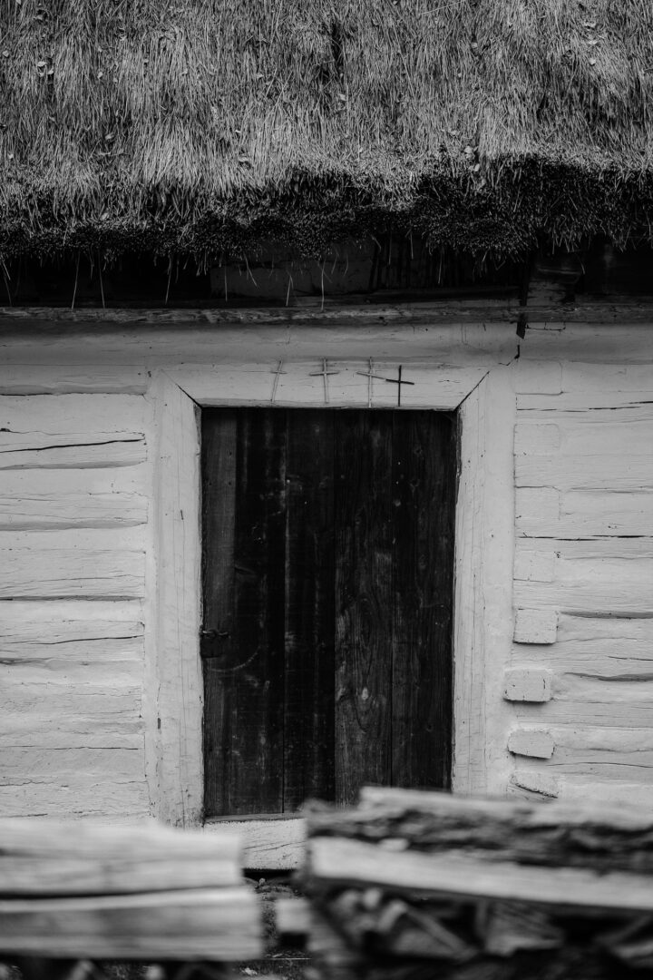 Weathered wooden door in a whitewashed log cabin with a thatched roof, small crosses above the doorway.