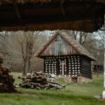 Traditional wooden log cabin with white circular patterns, thatched roof, set in a yard with stacked firewood.