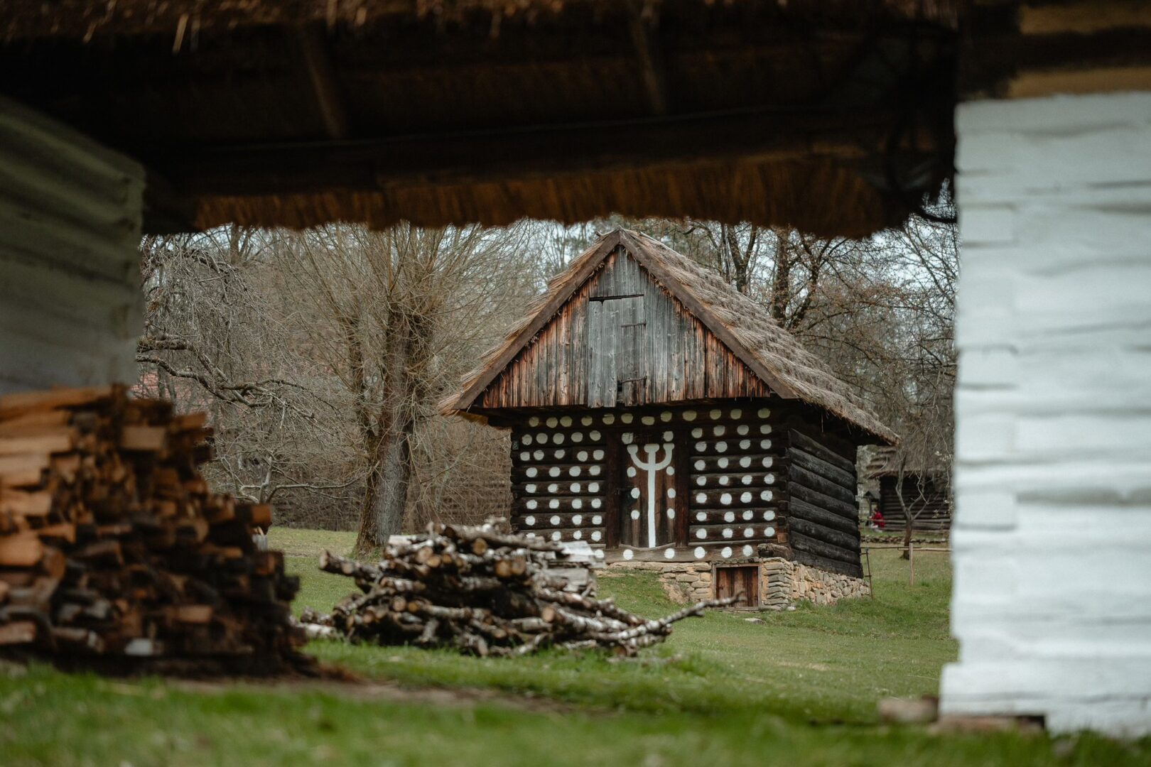 Traditional wooden log cabin with white circular patterns, thatched roof, set in a yard with stacked firewood.