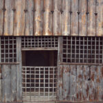 Weathered wooden facade with vertical planks, heart-shaped cutouts, and a central barred doorway like a rustic jail.