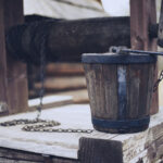 Old wooden bucket with metal bands sits on a weathered wooden platform, chained to a well mechanism, outdoors.