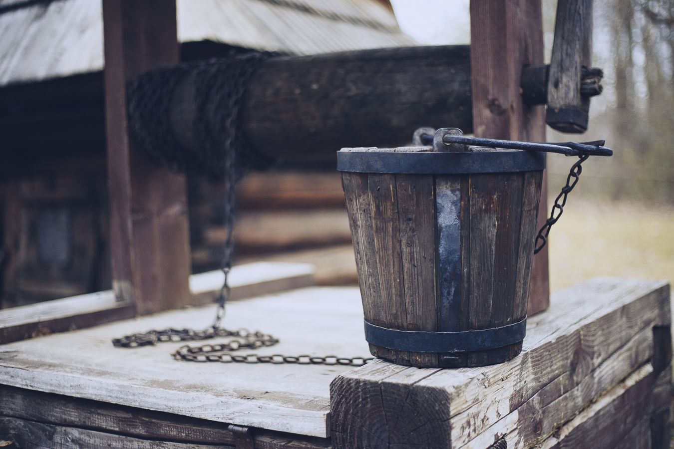 Old wooden bucket with metal bands sits on a weathered wooden platform, chained to a well mechanism, outdoors.