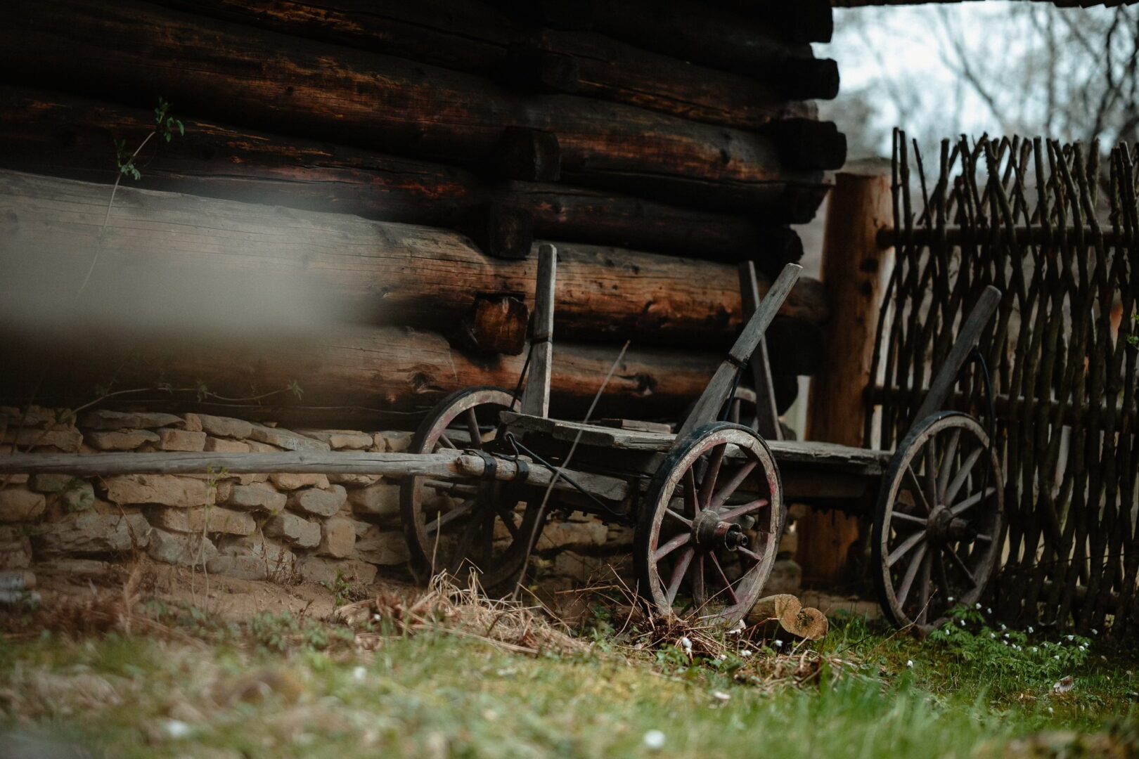 Old wooden wagon with spoked wheels resting beside a log cabin wall and rustic fence in a rural yard