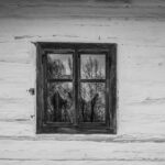 Weathered wooden wall with a small square window reflecting leafless trees in black and white.
