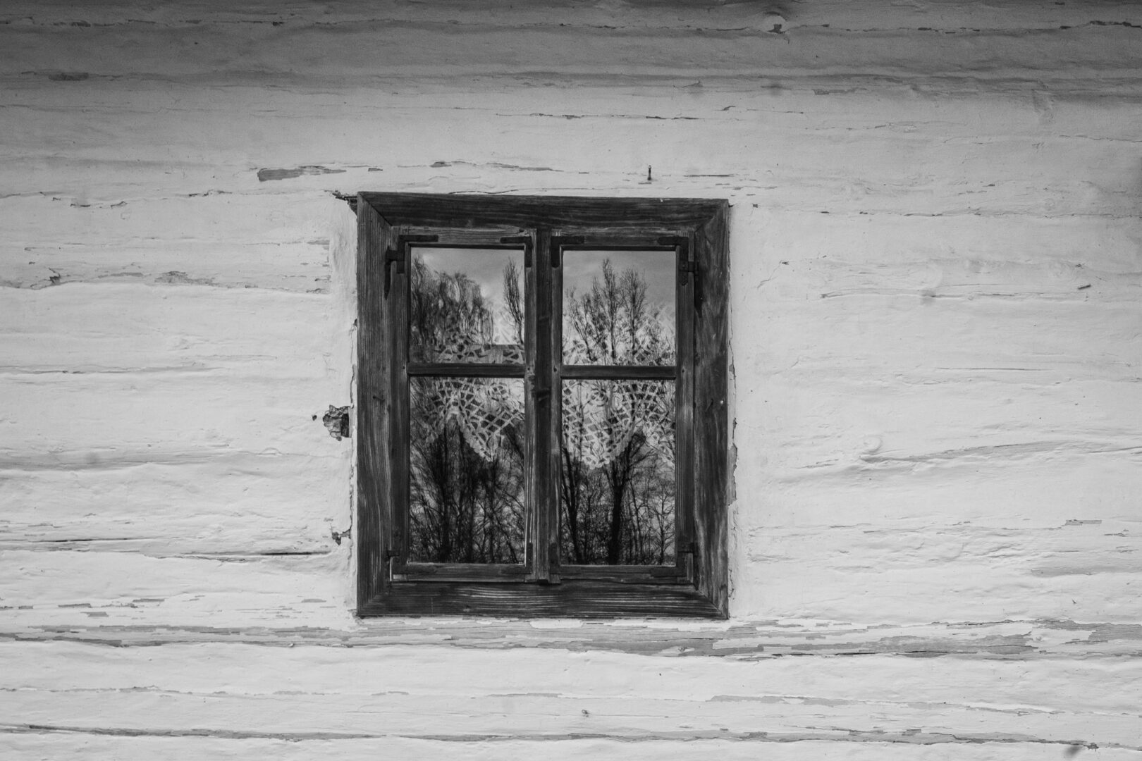 Weathered wooden wall with a small square window reflecting leafless trees in black and white.