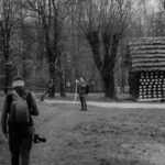 Photographer with a backpack approaches across a bare-tree park; a man in the distance is holding a long white scarf or ribbon near a wooden hut.