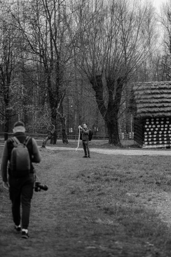 Photographer with a backpack approaches across a bare-tree park; a man in the distance is holding a long white scarf or ribbon near a wooden hut.