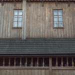 Weathered wooden house facade with two small-pane windows and a carved balustrade balcony below.