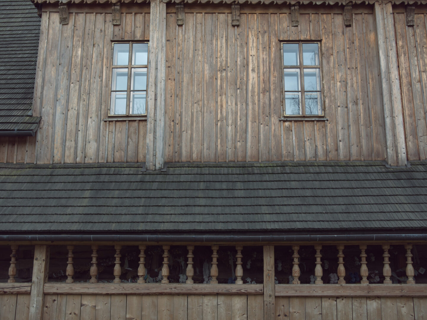 Weathered wooden house facade with two small-pane windows and a carved balustrade balcony below.