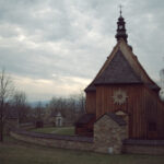 Wooden church with a tall steeple and cross, surrounded by a stone wall and leafless trees on a cloudy day.