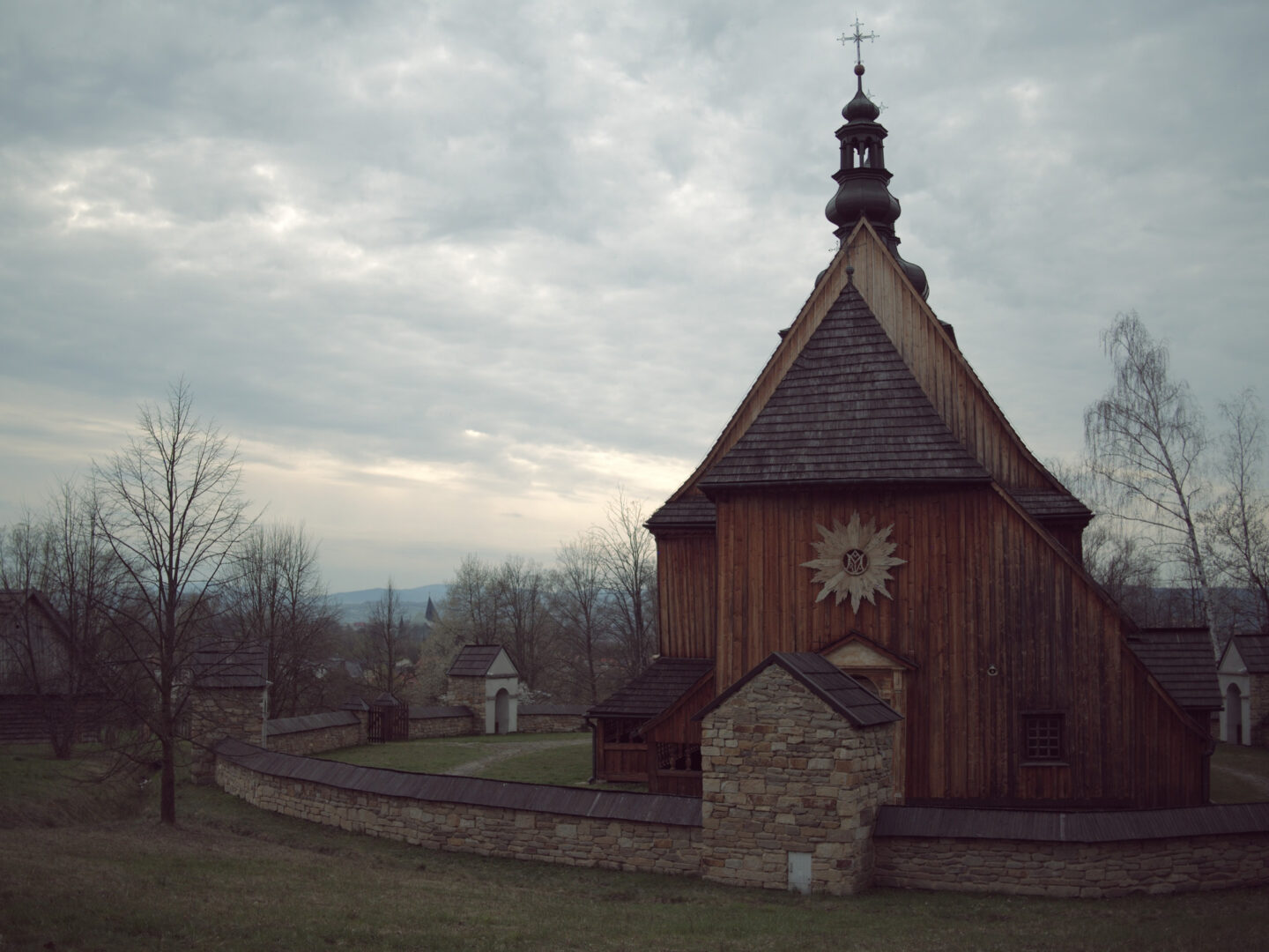 Wooden church with a tall steeple and cross, surrounded by a stone wall and leafless trees on a cloudy day.