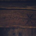 Carved circular emblem with an X-like four-pointed star inside on a dark, weathered wooden beam, with faded numbers to the left.