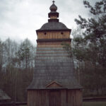 Tall wooden church tower with a tiered shingle roof in a forest under an overcast sky, cross atop the steeple.