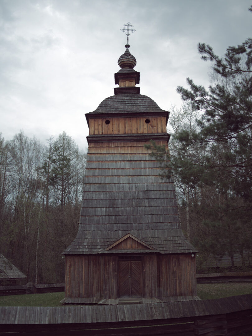 Tall wooden church tower with a tiered shingle roof in a forest under an overcast sky, cross atop the steeple.