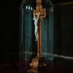 Ornate crucifix with Jesus statue under a glass dome, on a gilded stand in a dim setting