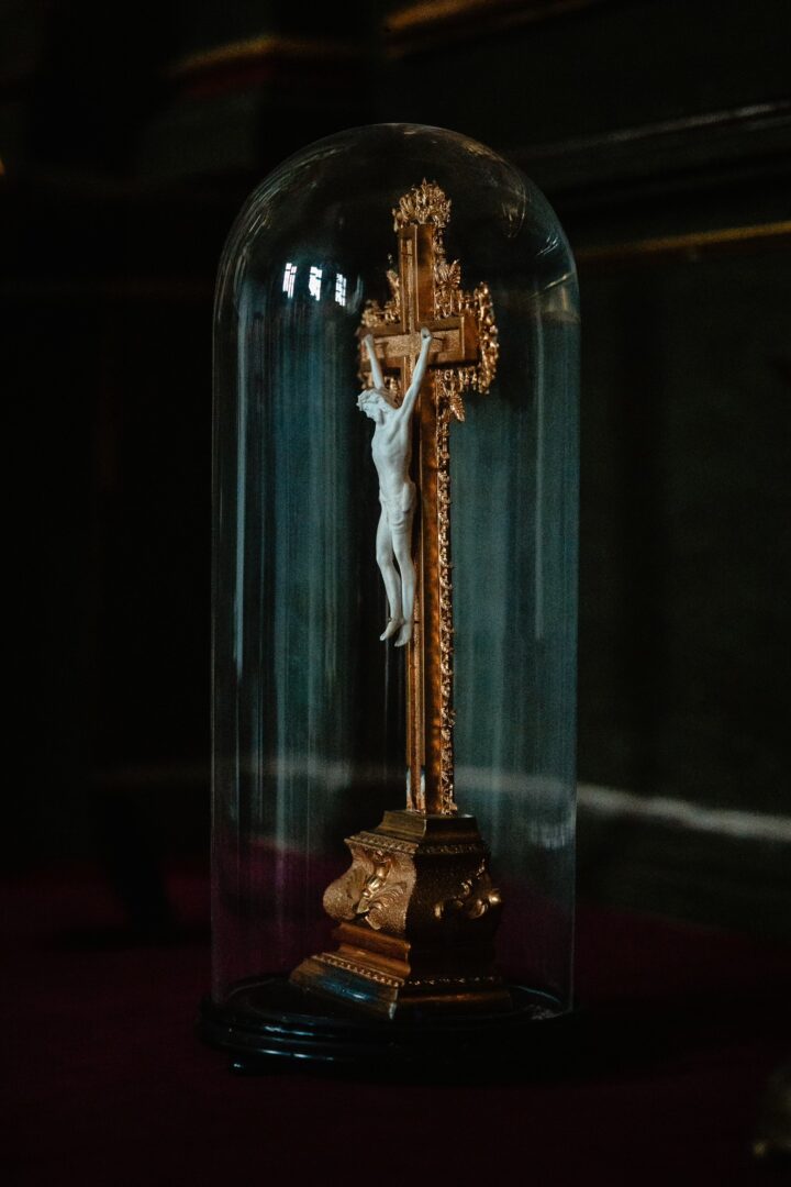 Ornate crucifix with Jesus statue under a glass dome, on a gilded stand in a dim setting