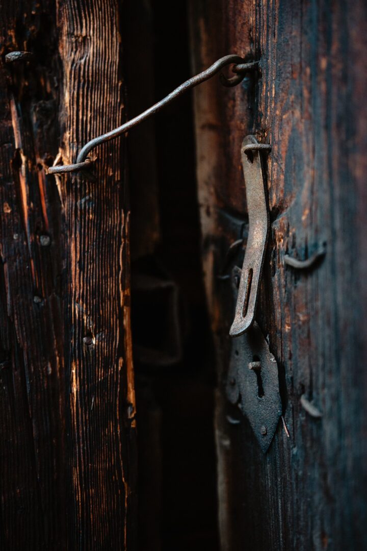 Old wooden door with a rusted latch and bar in a dim, narrow passageway behind it.