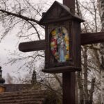 Rustic wooden roadside shrine with a flower-framed statue of Mary mounted on a post among bare trees outside.