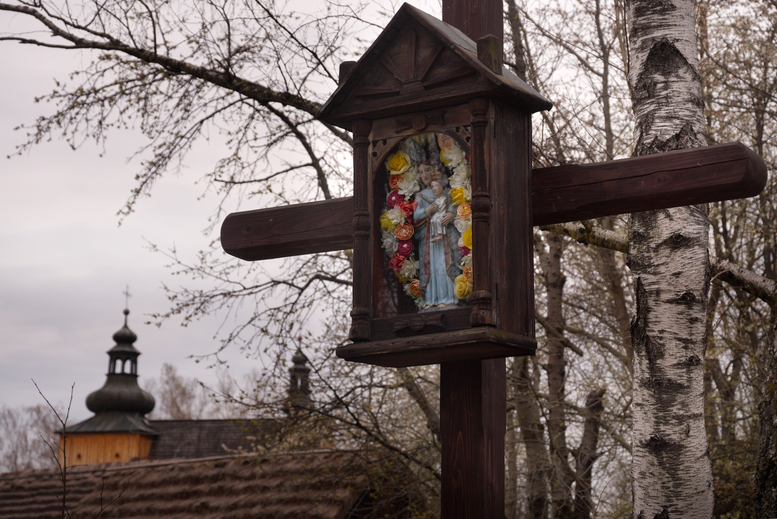 Rustic wooden roadside shrine with a flower-framed statue of Mary mounted on a post among bare trees outside.