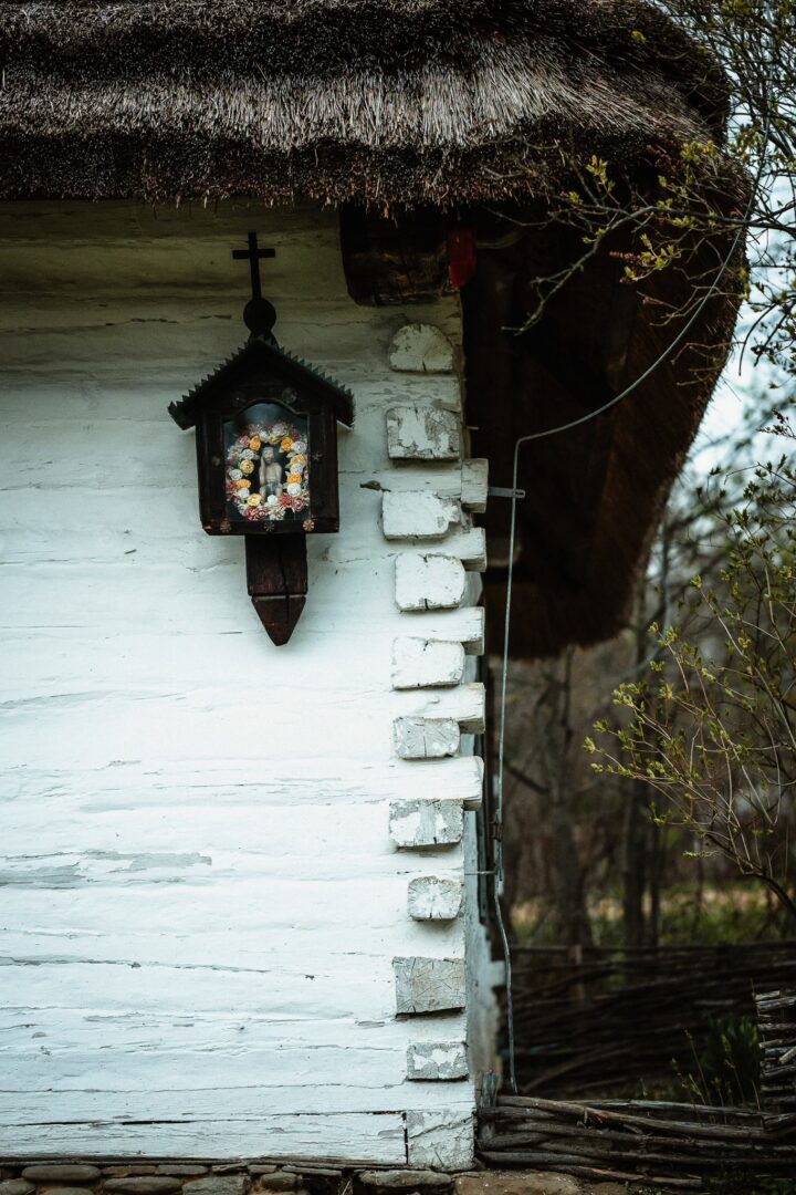 Outdoor wooden shrine with a crucifix mounted on a white-washed wall of a thatched-roof cottage.