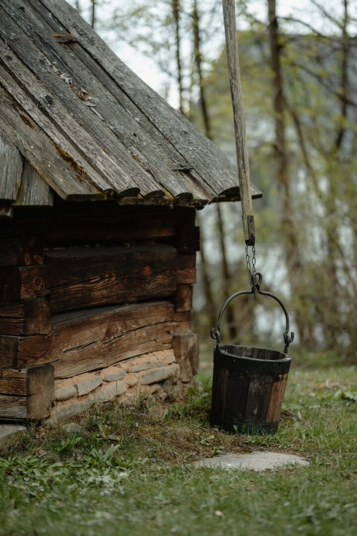 Rustic log cabin with a weathered wooden bucket hanging from a rope beside it in a forest clearing