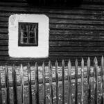 Black-and-white photo of a rustic wooden house with a small square window framed in white plaster, and a wooden picket fence in front.