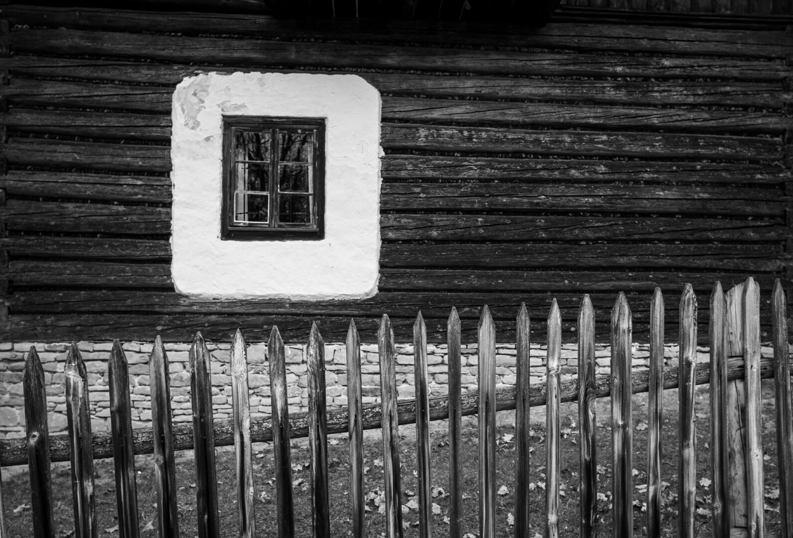 Black-and-white photo of a rustic wooden house with a small square window framed in white plaster, and a wooden picket fence in front.
