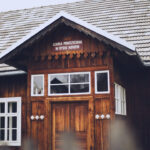 Front of a traditional wooden schoolhouse with white-framed windows and a gabled roof, a Polish sign above the door reads SZKOŁA POWSZECHNA W RYBIU NOWEM, set against a clear sky.