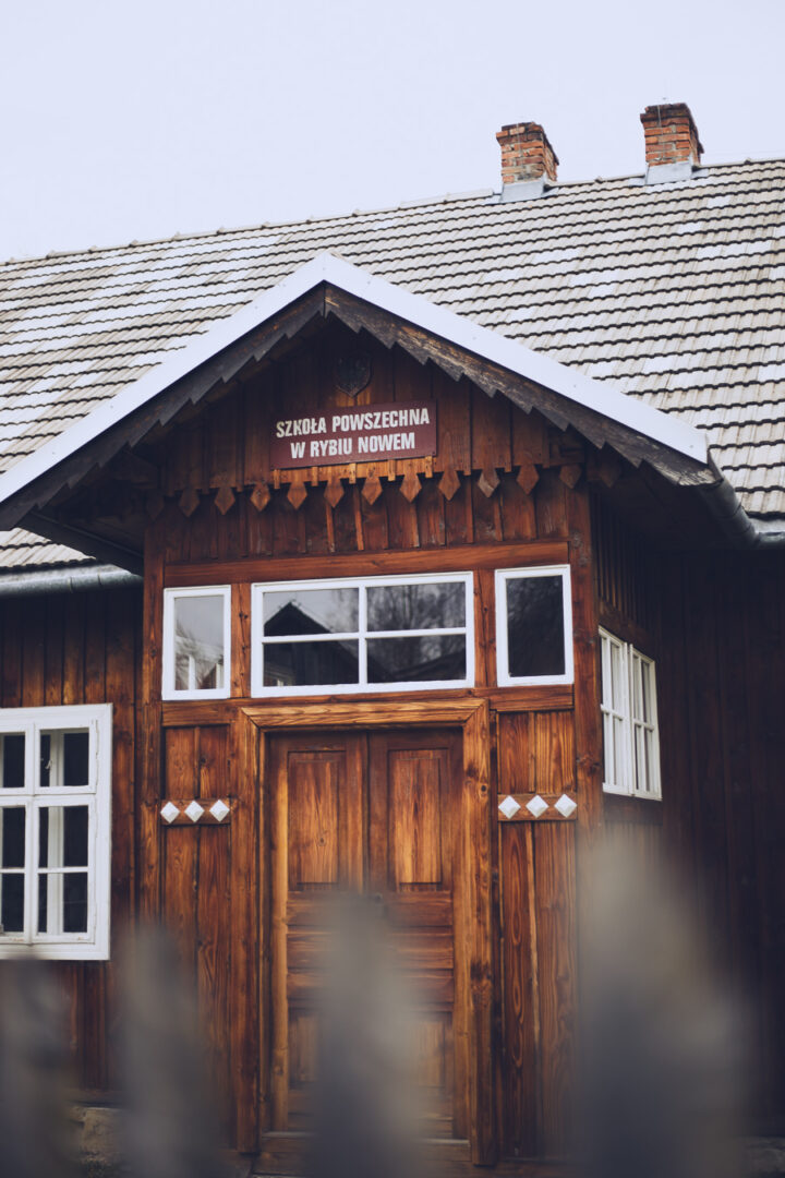 Front of a traditional wooden schoolhouse with white-framed windows and a gabled roof, a Polish sign above the door reads SZKOŁA POWSZECHNA W RYBIU NOWEM, set against a clear sky.