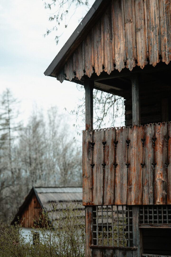 Weathered wooden cabin with carved balcony railing in a rural, leafless landscape.