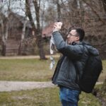 Man in a gray jacket and glasses holds a pink and white paper garland up toward bare tree branches in a park.