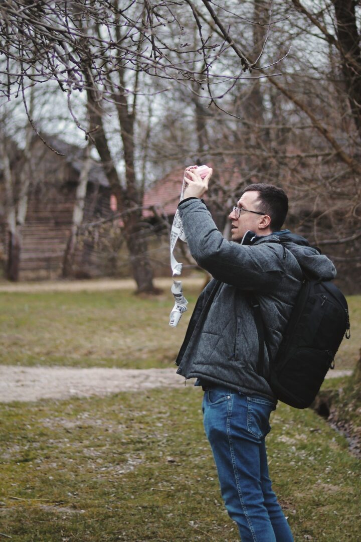 Man in a gray jacket and glasses holds a pink and white paper garland up toward bare tree branches in a park.