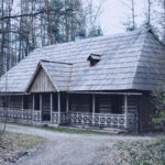 Rustic wooden cabin in a leafless forest, featuring a wraparound porch and a steep shingled roof.