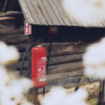 Red fire extinguisher mounted on a wooden post beside a rustic log cabin, with blurred white flowers in the foreground.