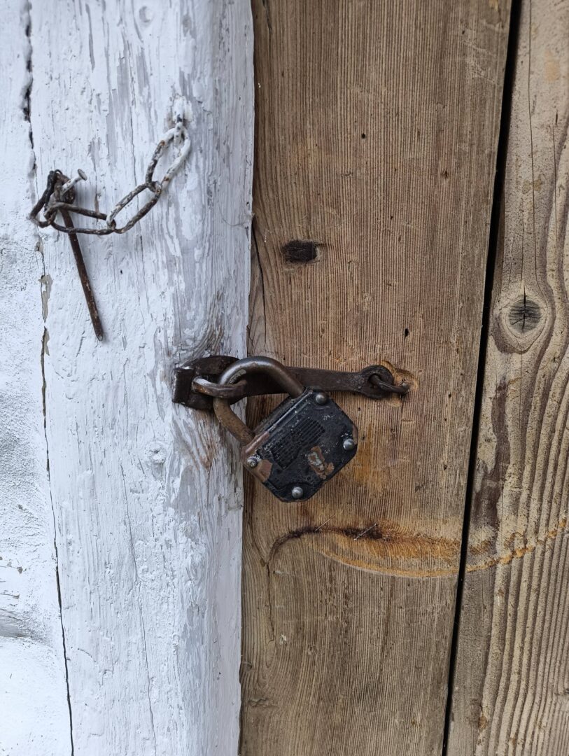 Rusty padlock and chain securing a weathered wooden door with peeling white paint on the left panel.