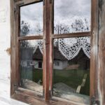 Old wooden window with lace curtains; view of a rural yard and a small white house outside.