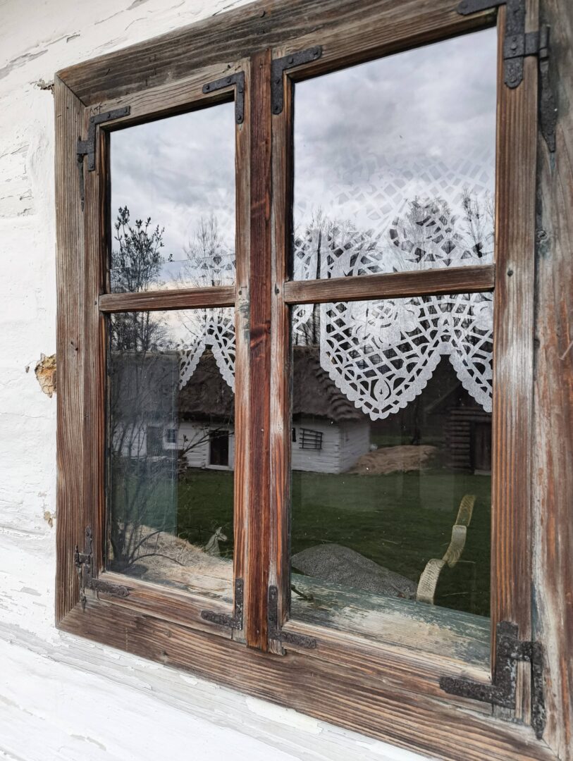 Old wooden window with lace curtains; view of a rural yard and a small white house outside.