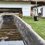 Weathered stone trough filled with still water in a rural farmyard; white thatched buildings rise behind the scene.
