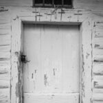 Weathered wooden door in a peeling white wall, with a row of metal crosses mounted above it and a small window high above.