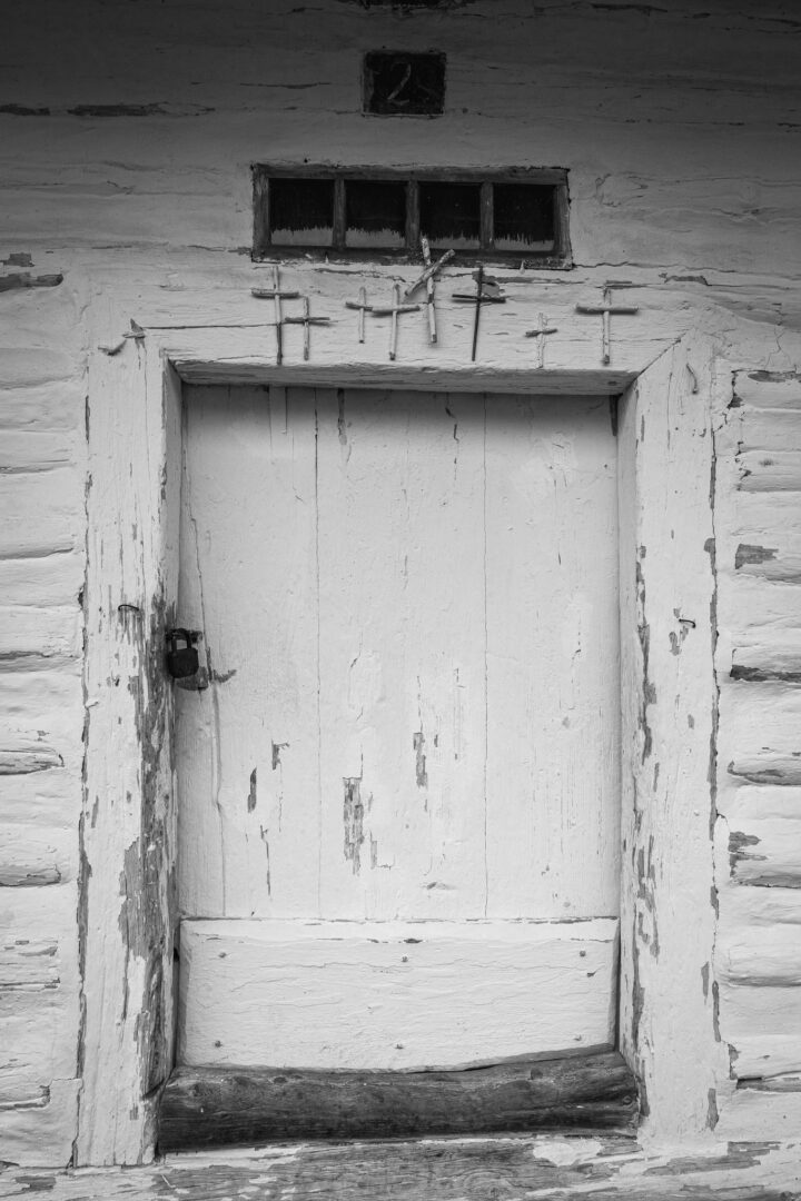 Weathered wooden door in a peeling white wall, with a row of metal crosses mounted above it and a small window high above.