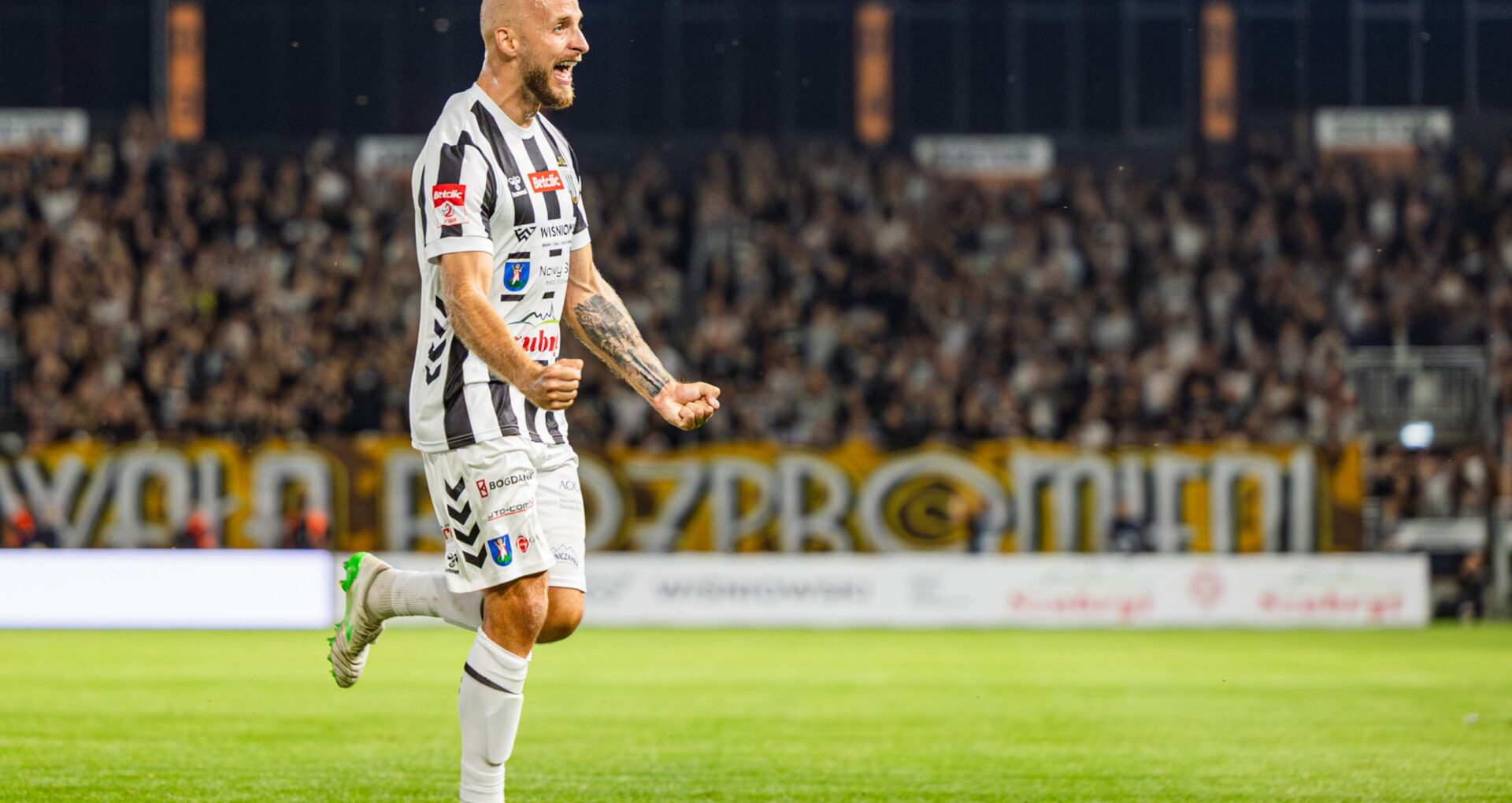 Soccer player in black-and-white striped kit celebrates on the field, fist clenched, crowd cheering in the stands behind him.