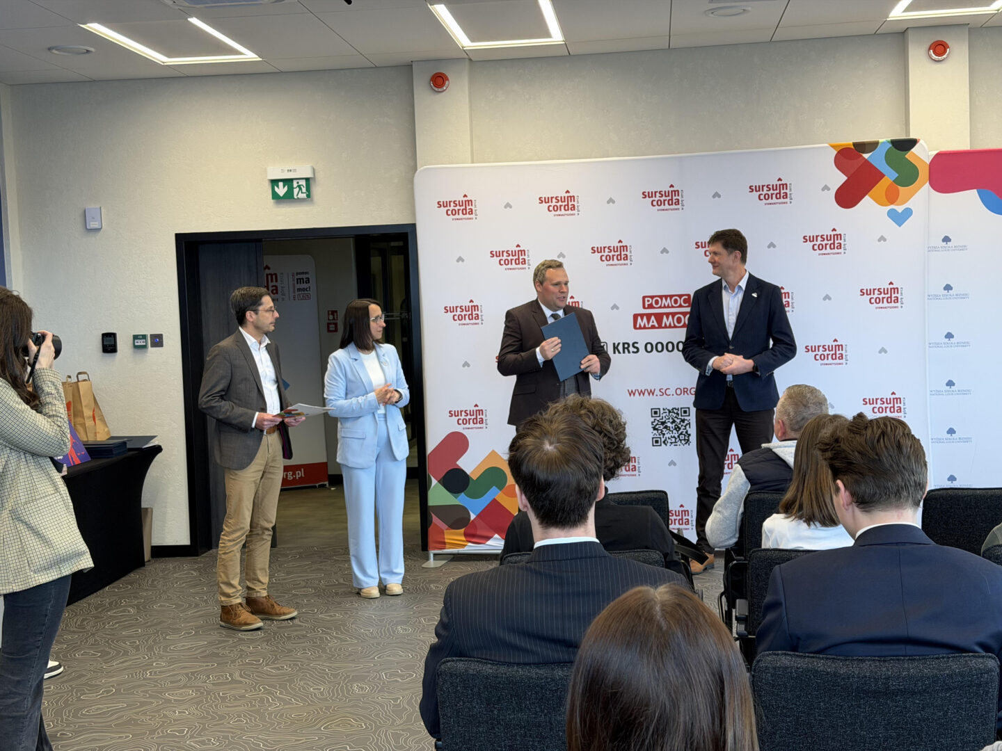 Three speakers stand at a stage beside a branded backdrop while an audience sits facing them; a photographer takes a picture on the left.