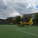 Two yellow air ambulance helicopters parked on a green sports field with a team in red near the left edge under a cloudy sky.