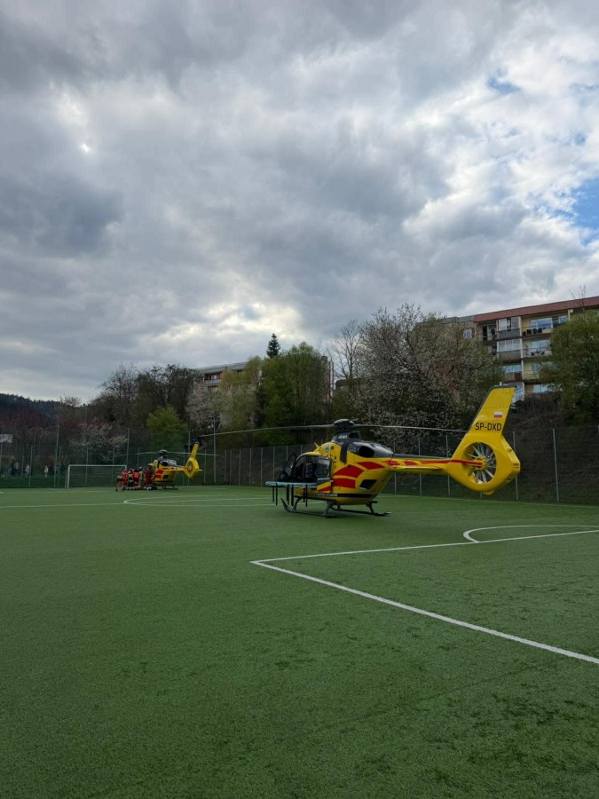 Two yellow air ambulance helicopters parked on a green sports field with a team in red near the left edge under a cloudy sky.