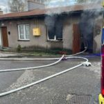 Residential street with a single-story house emitting smoke from a doorway; fire hoses on wet pavement lead to a nearby fire truck.