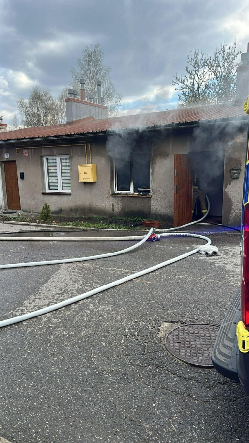Residential street with a single-story house emitting smoke from a doorway; fire hoses on wet pavement lead to a nearby fire truck.