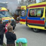 Emergency scene with three yellow and red ambulances, smoke rising from a building, and bystanders watching.