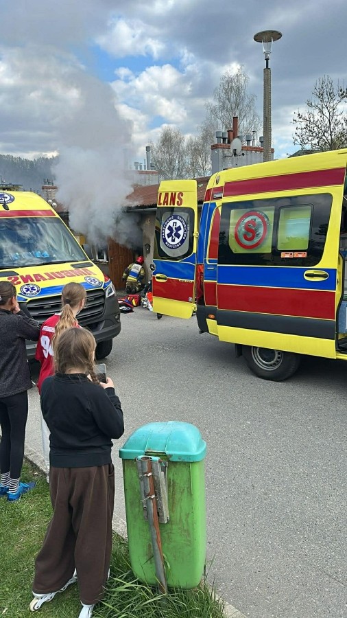 Emergency scene with three yellow and red ambulances, smoke rising from a building, and bystanders watching.