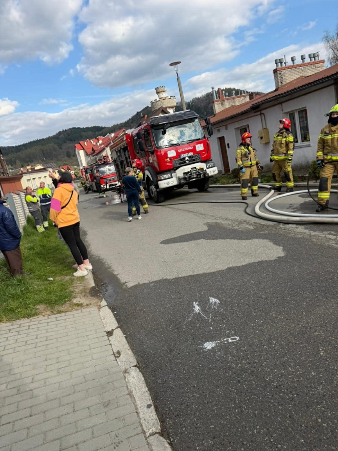 Firefighters in turnout gear with a red fire engine on a residential street, hoses laid out and onlookers nearby.