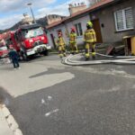 Firefighters in yellow turnout gear standing on a residential street with a red fire truck and coiled hoses nearby.